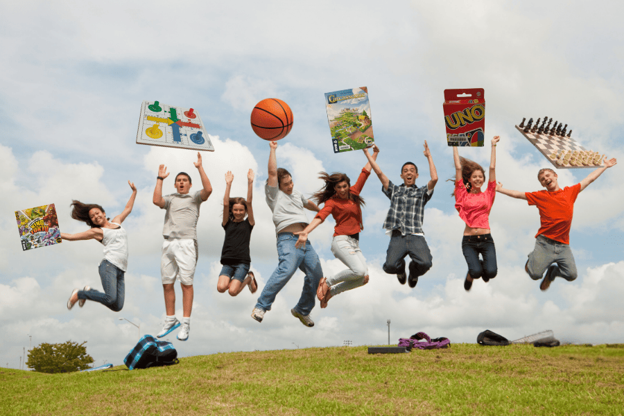 Young people jumping with board games like Parcheesi, UNO, chess, and a basketball. Festive, fun atmosphere outdoors.